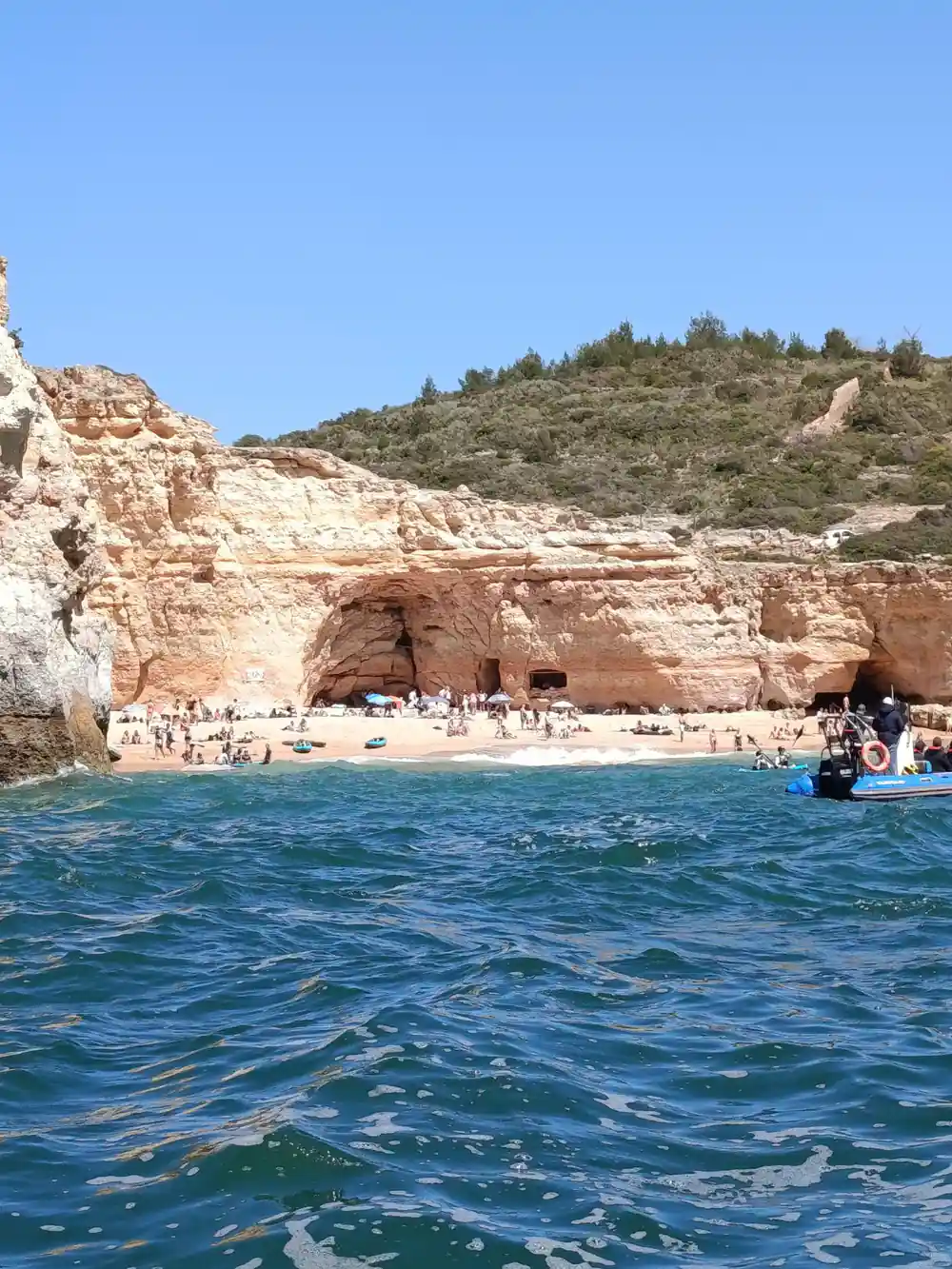 A crowded beach beneath layered cliffs with caves at the back